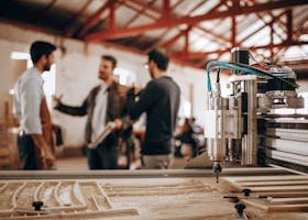 Three men discussing work in a woodworking shop with CNC machinery in focus.